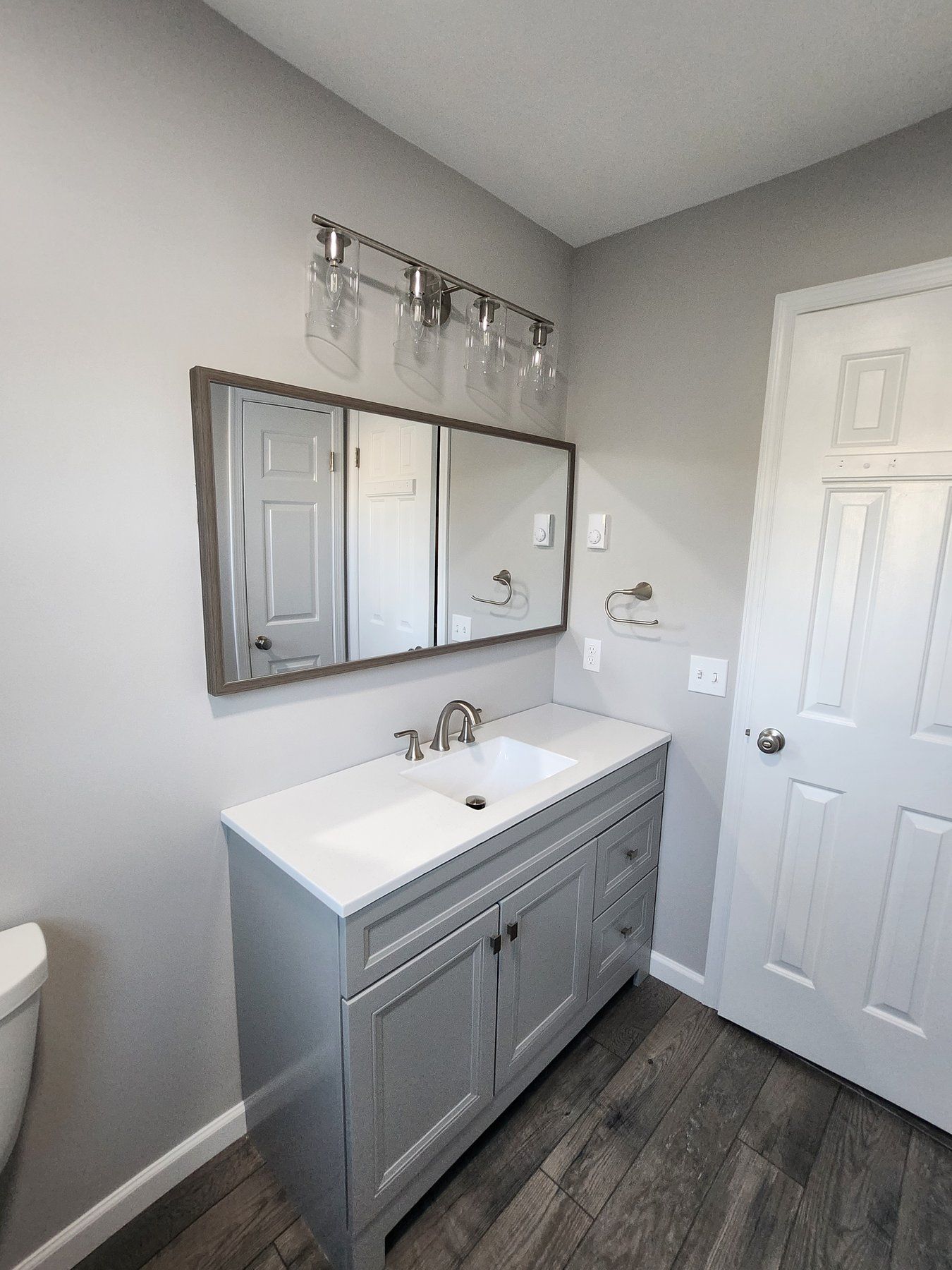 Small bathroom with gray vanity, white sink, mirror, and white door on wood-look flooring.
