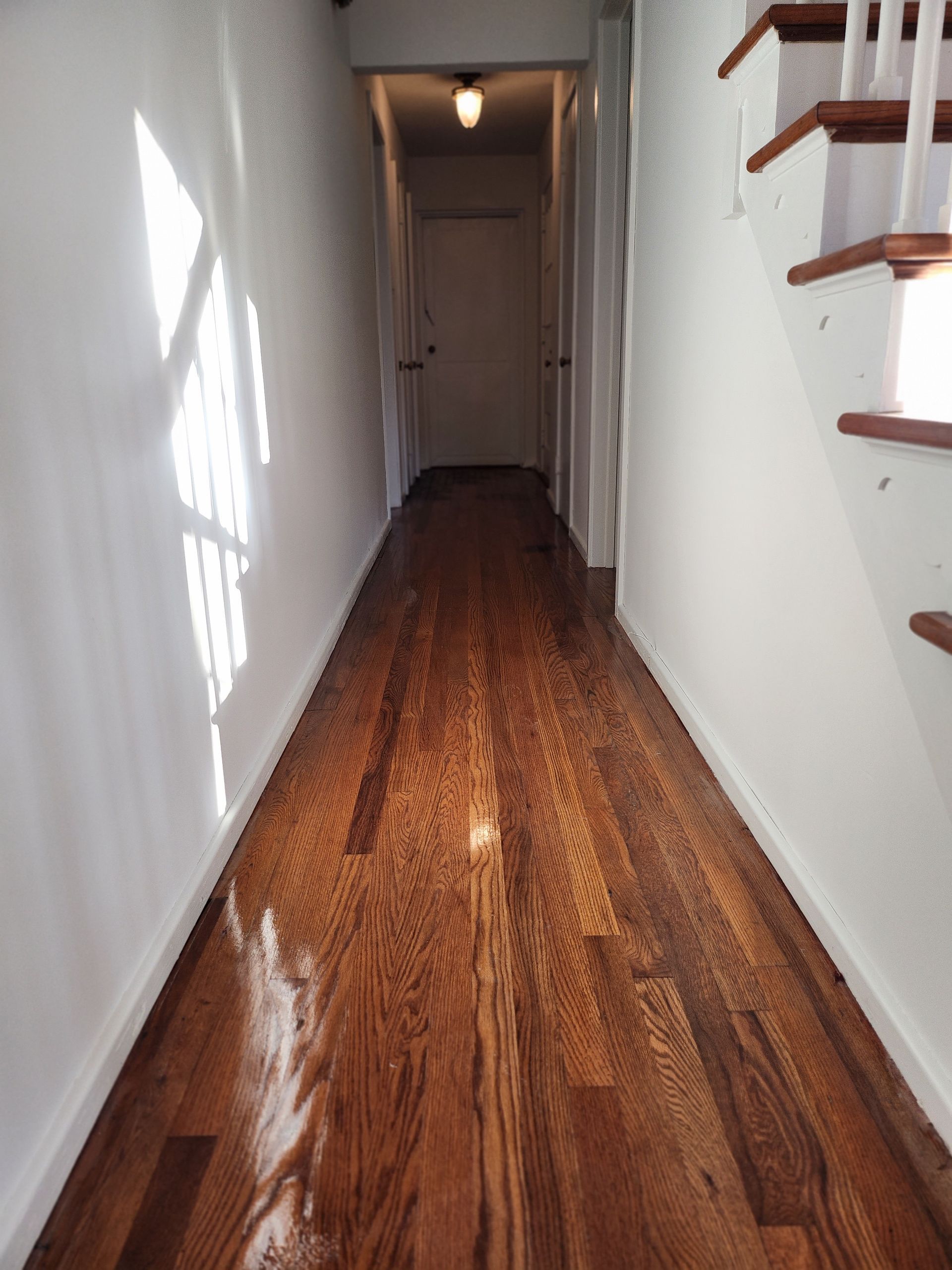 Narrow hallway with polished hardwood floor, white walls, and staircase railing on the right.