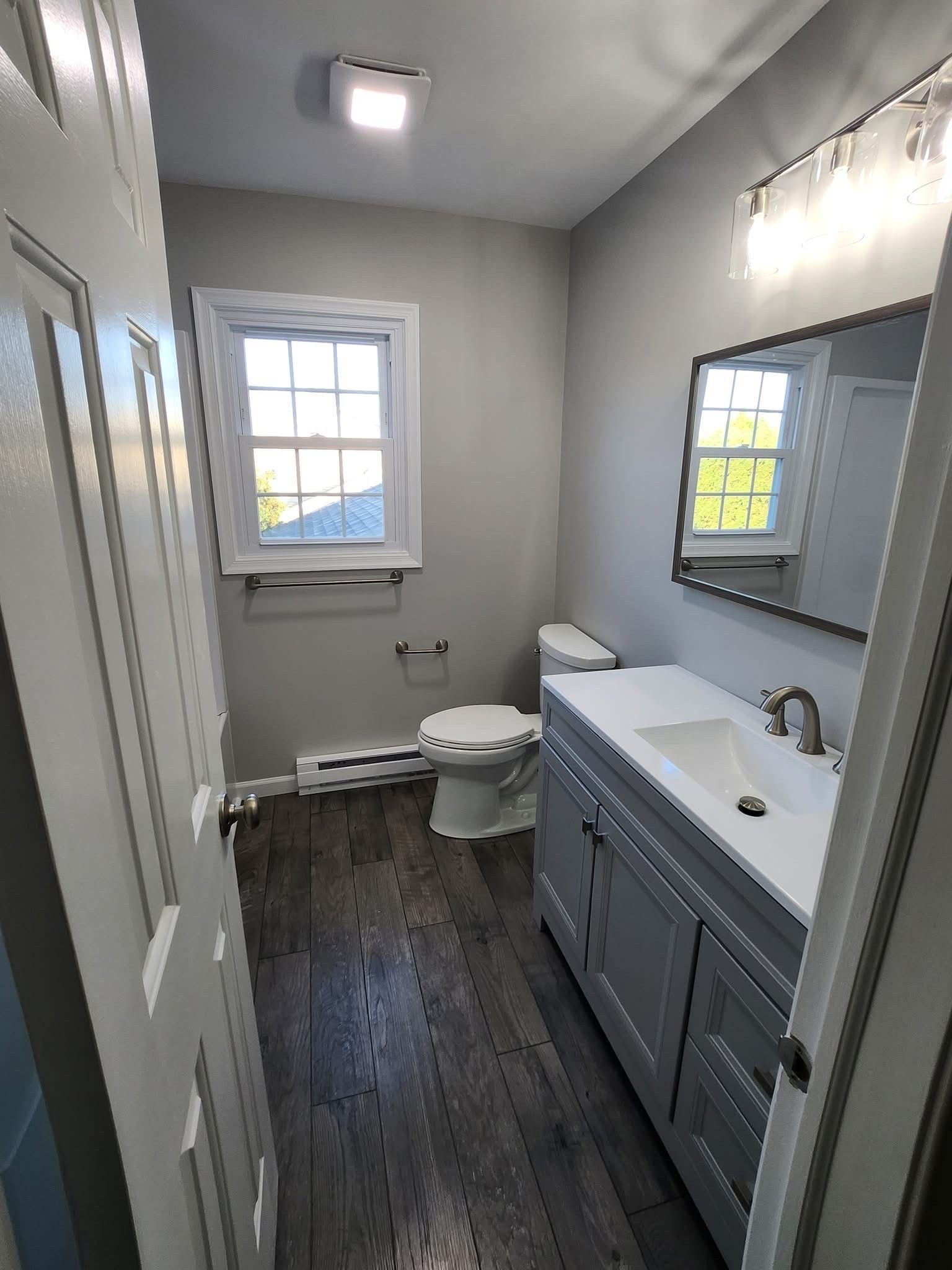Narrow modern bathroom with gray vanity, white toilet, window, mirror, and dark wood floor.