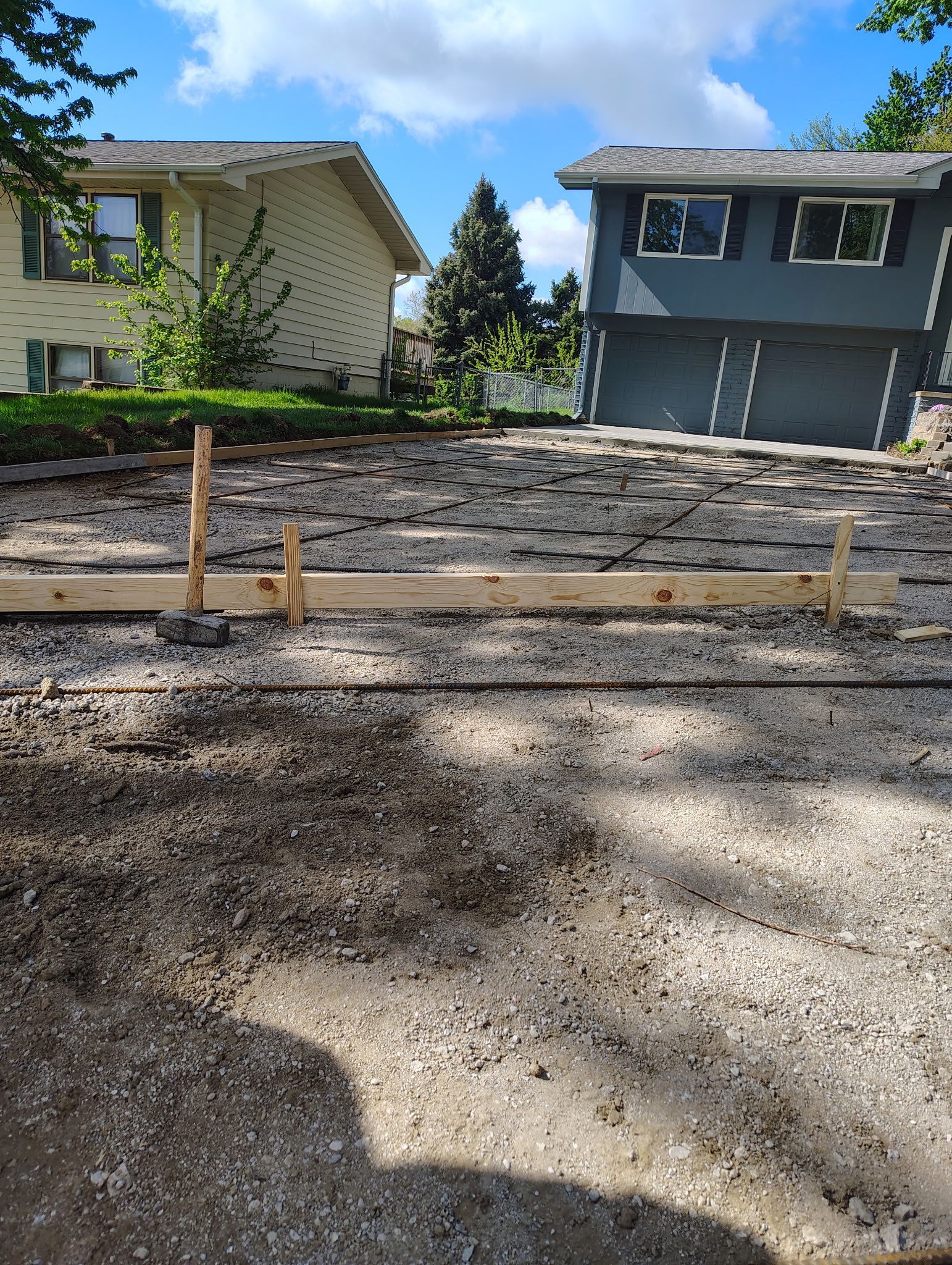 Front yard construction site with gravel, wooden stakes, and two houses under a blue sky
