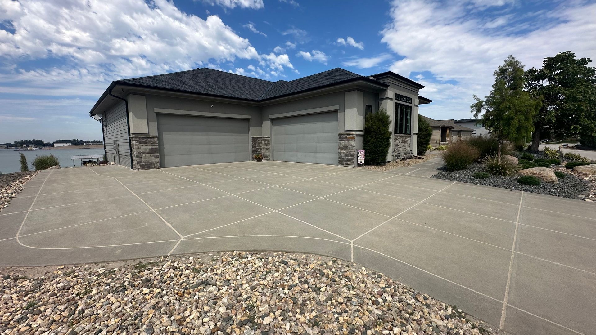 Modern gray house with a three-car garage, large concrete driveway, and rock landscaping under a cloudy sky