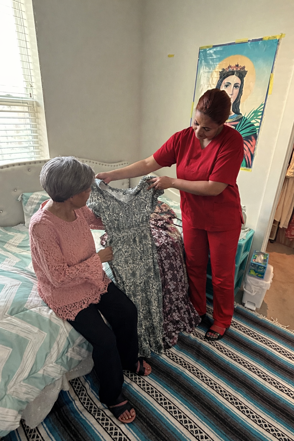 Woman in red scrubs shows dress to a seated person. Bedroom setting; bed, rug, painting on wall.