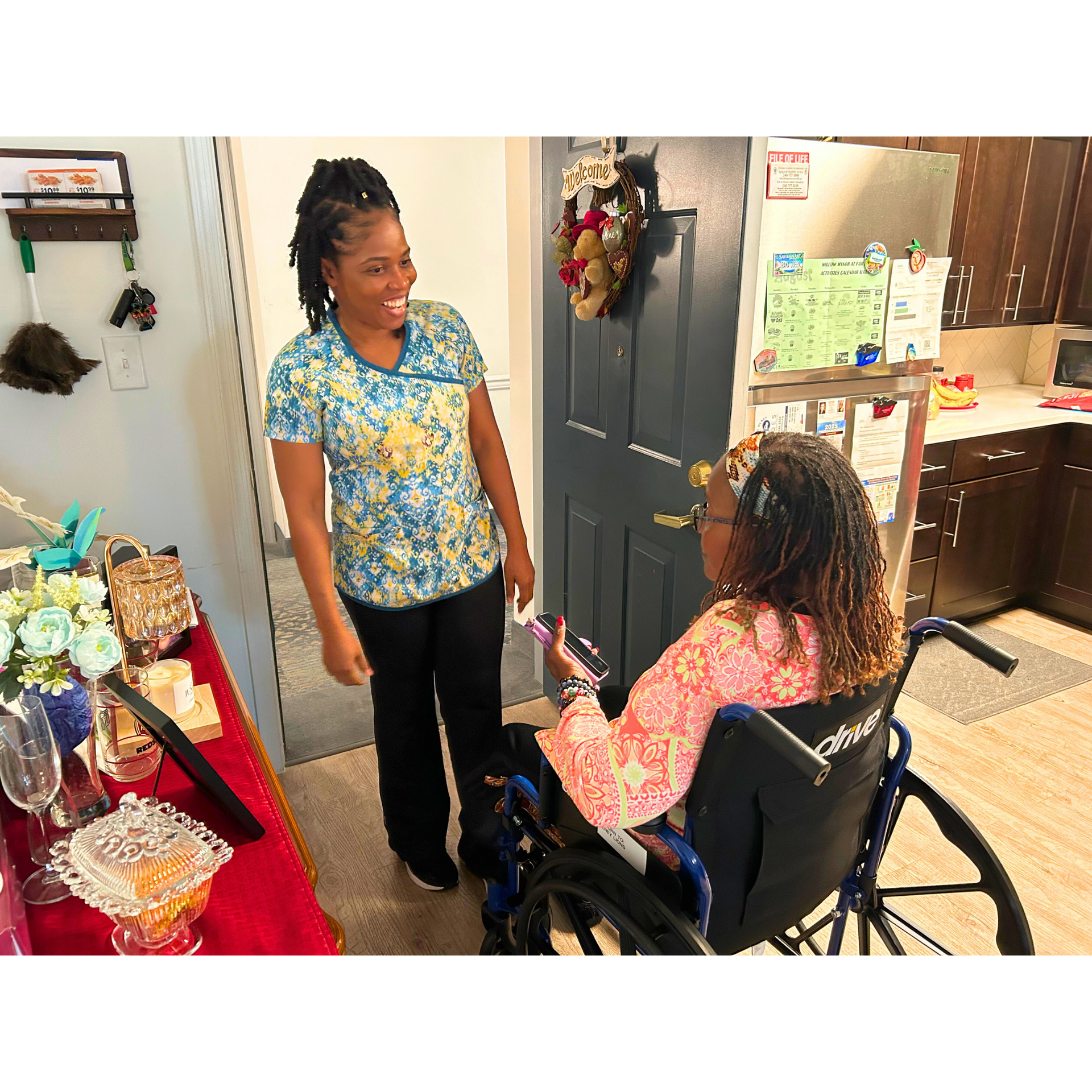 Woman in blue floral top smiles at woman in wheelchair near doorway with gifts on a table.