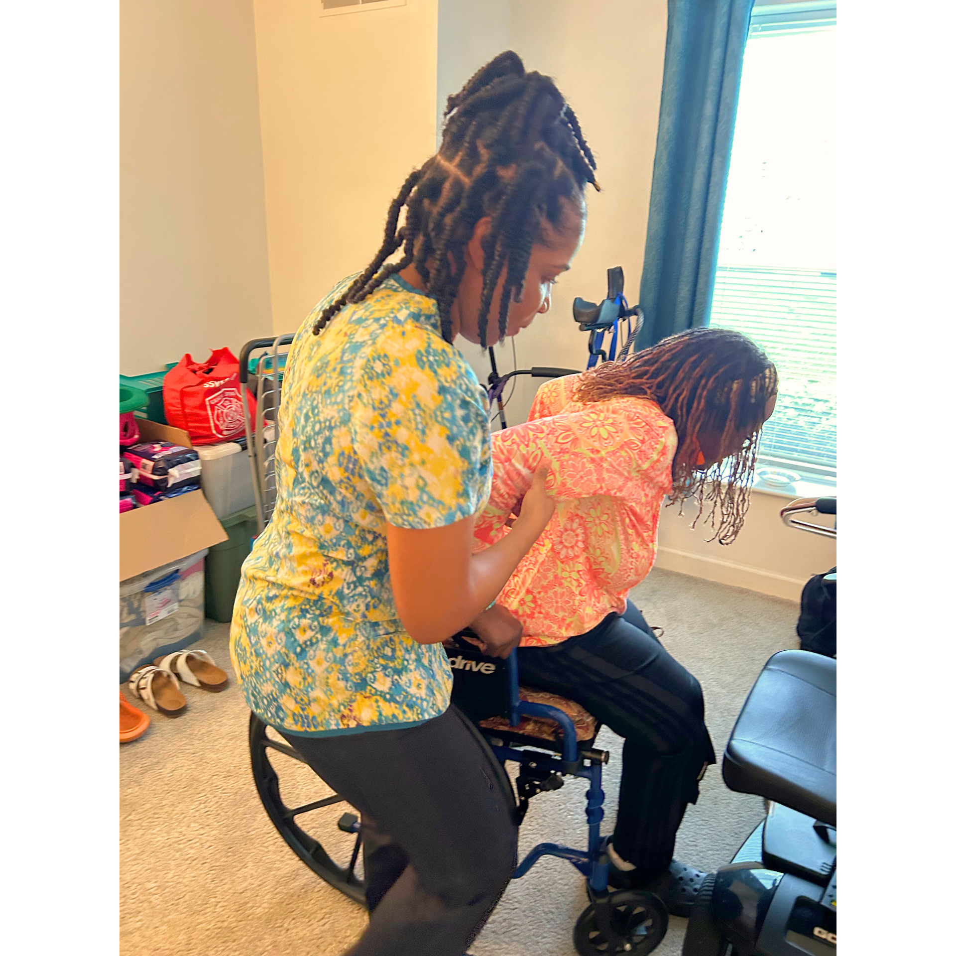 Woman assisting another person from a wheelchair, indoors. They both have braided hair and wear casual clothing.