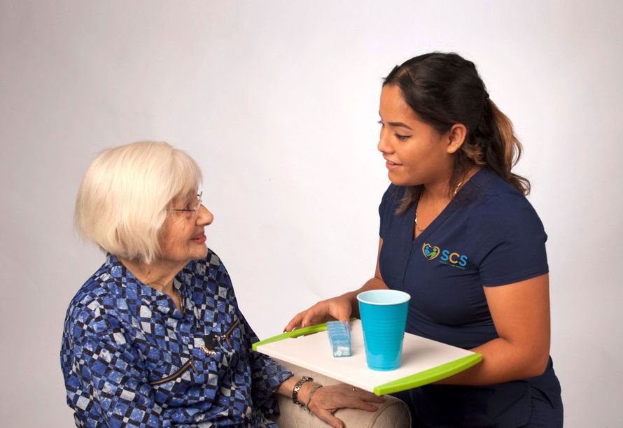 A caregiver offers medication and water to an elderly person in a room with a white background.