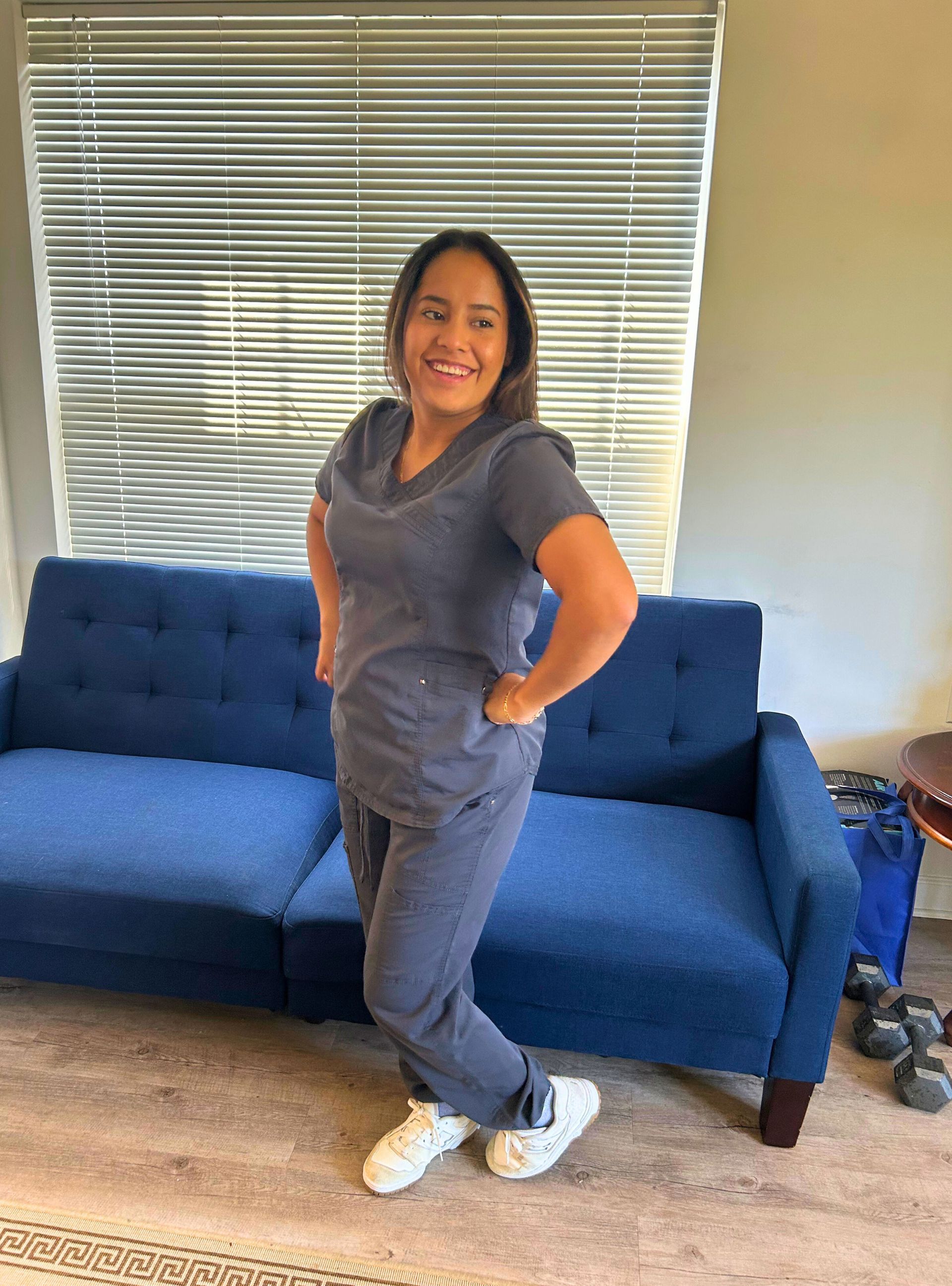 Woman in blue scrubs smiles, posing with hands on hips, near a blue sofa and window blinds.