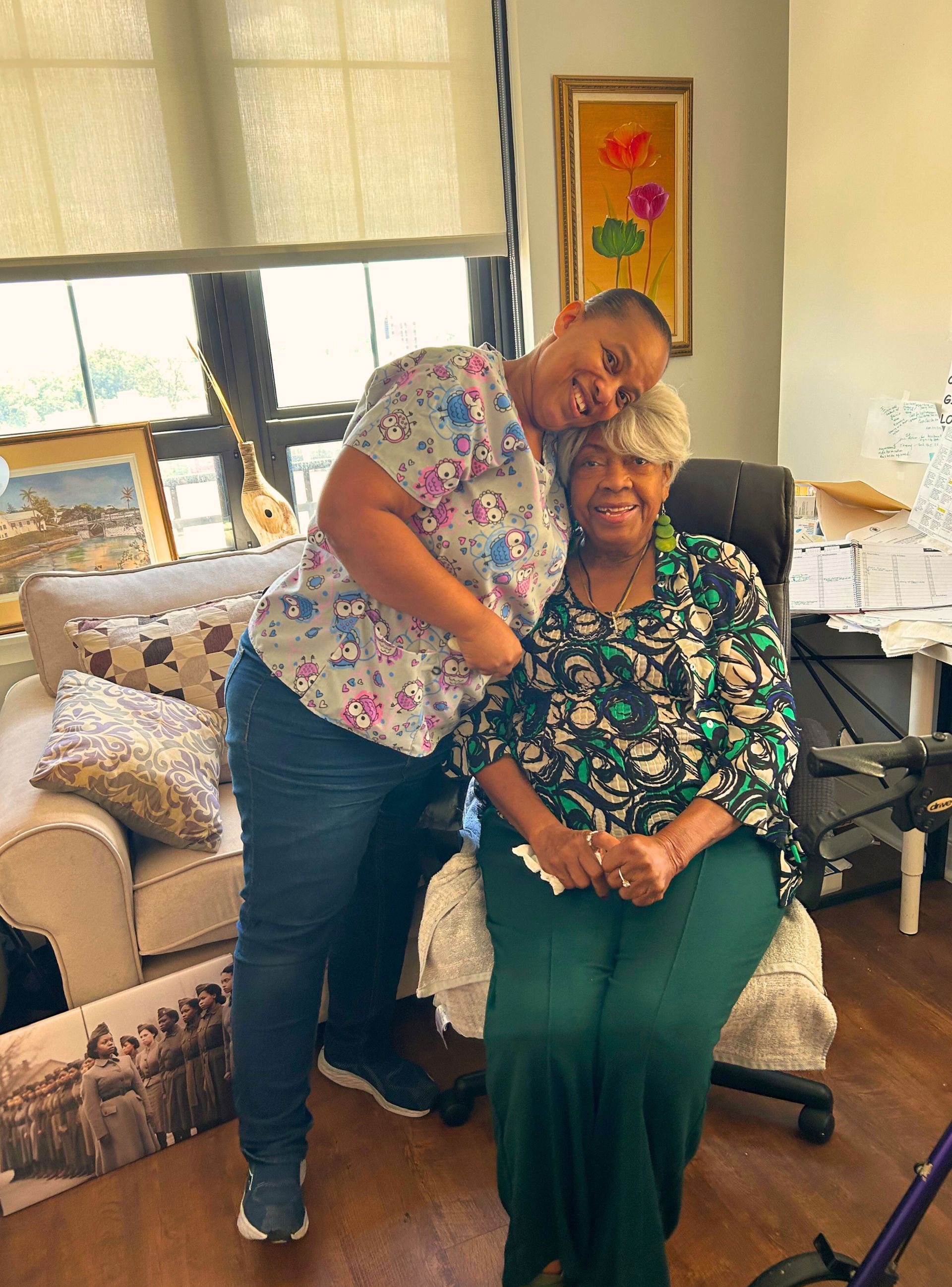 Woman in floral top hugs seated woman in green pants; smiles indoors.