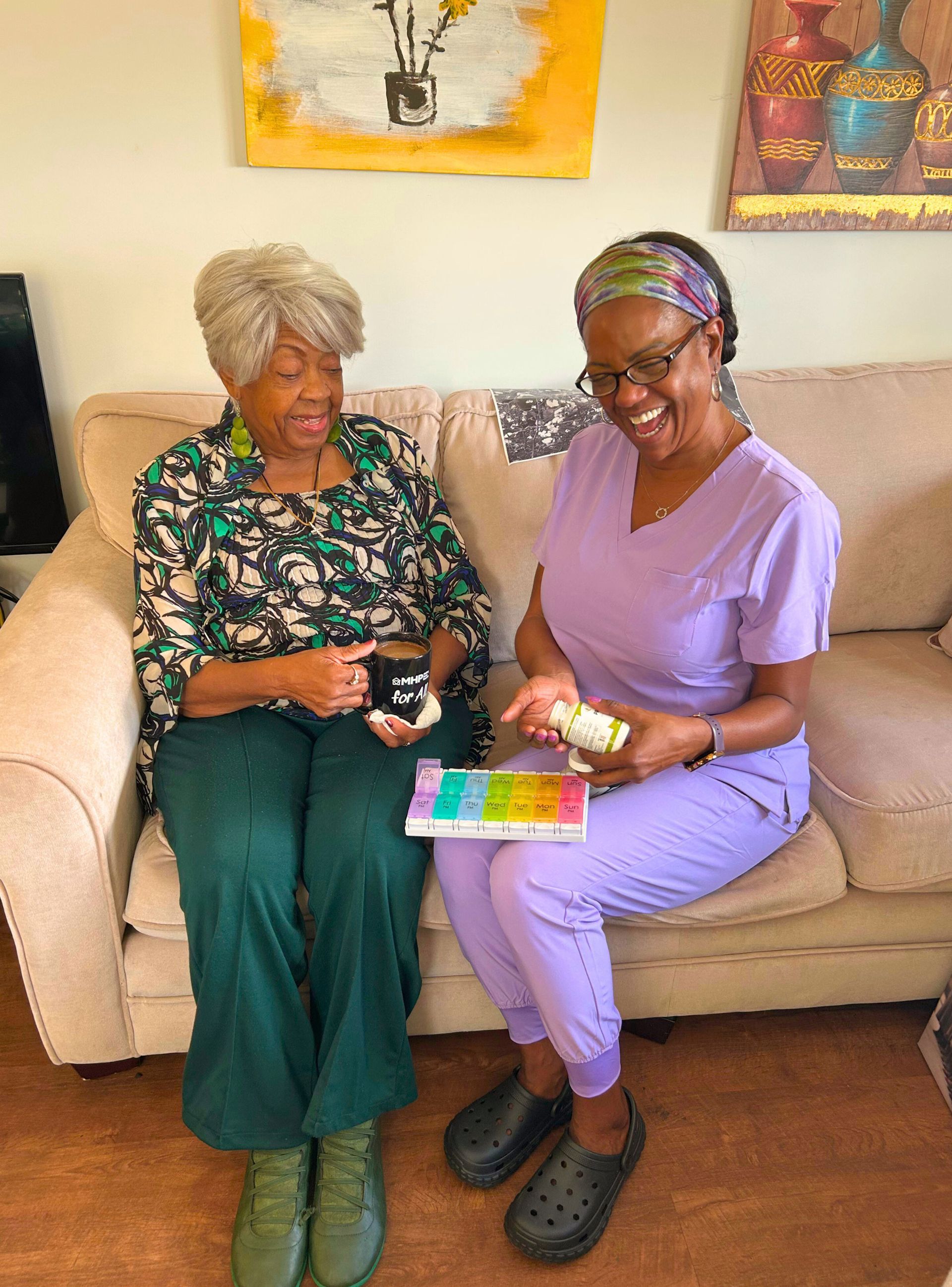 Woman showing medication to another woman on a couch; indoor setting. Both smiling.