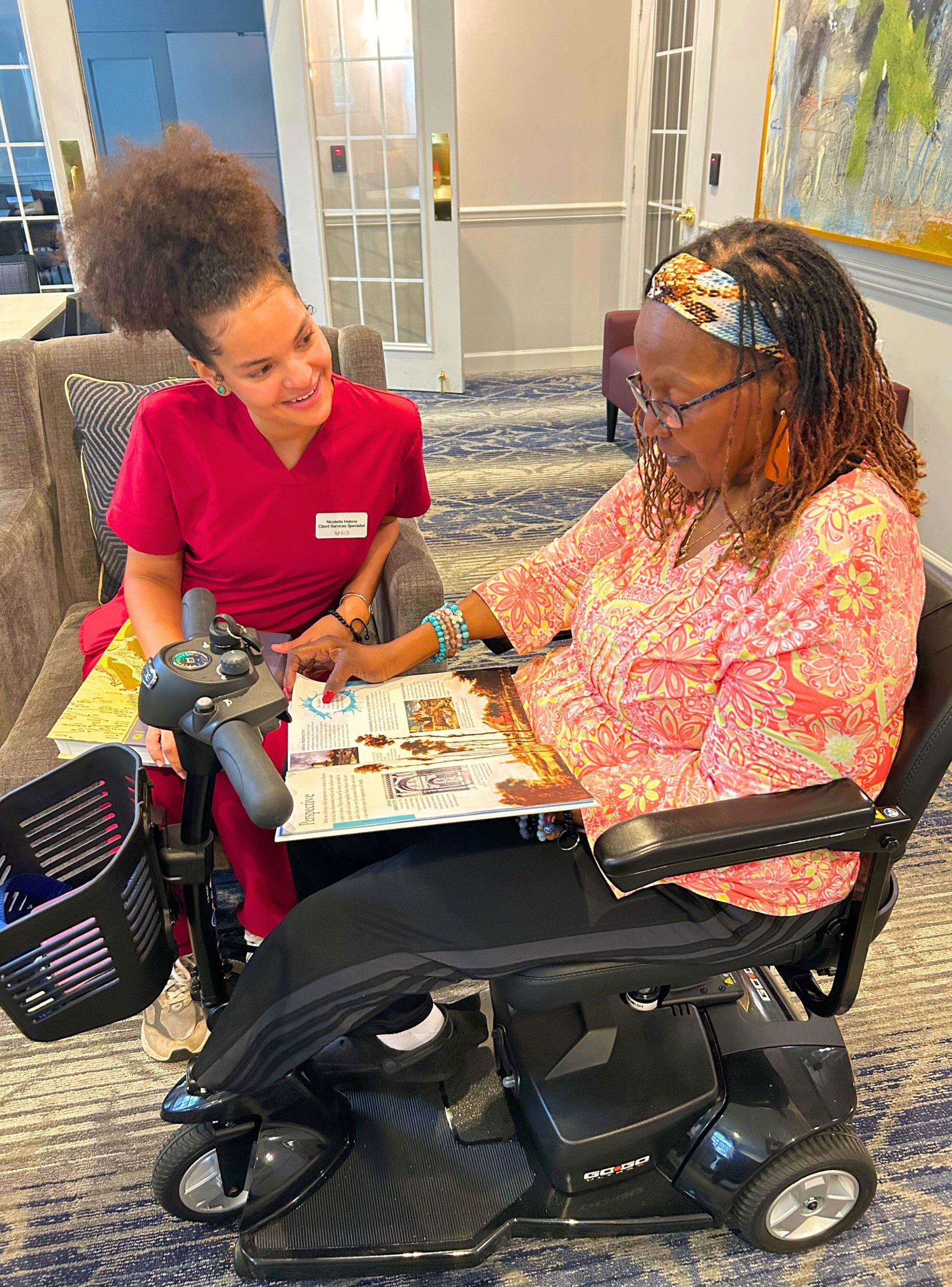 Caregiver shows magazine to person in wheelchair, both smiling, indoor setting.