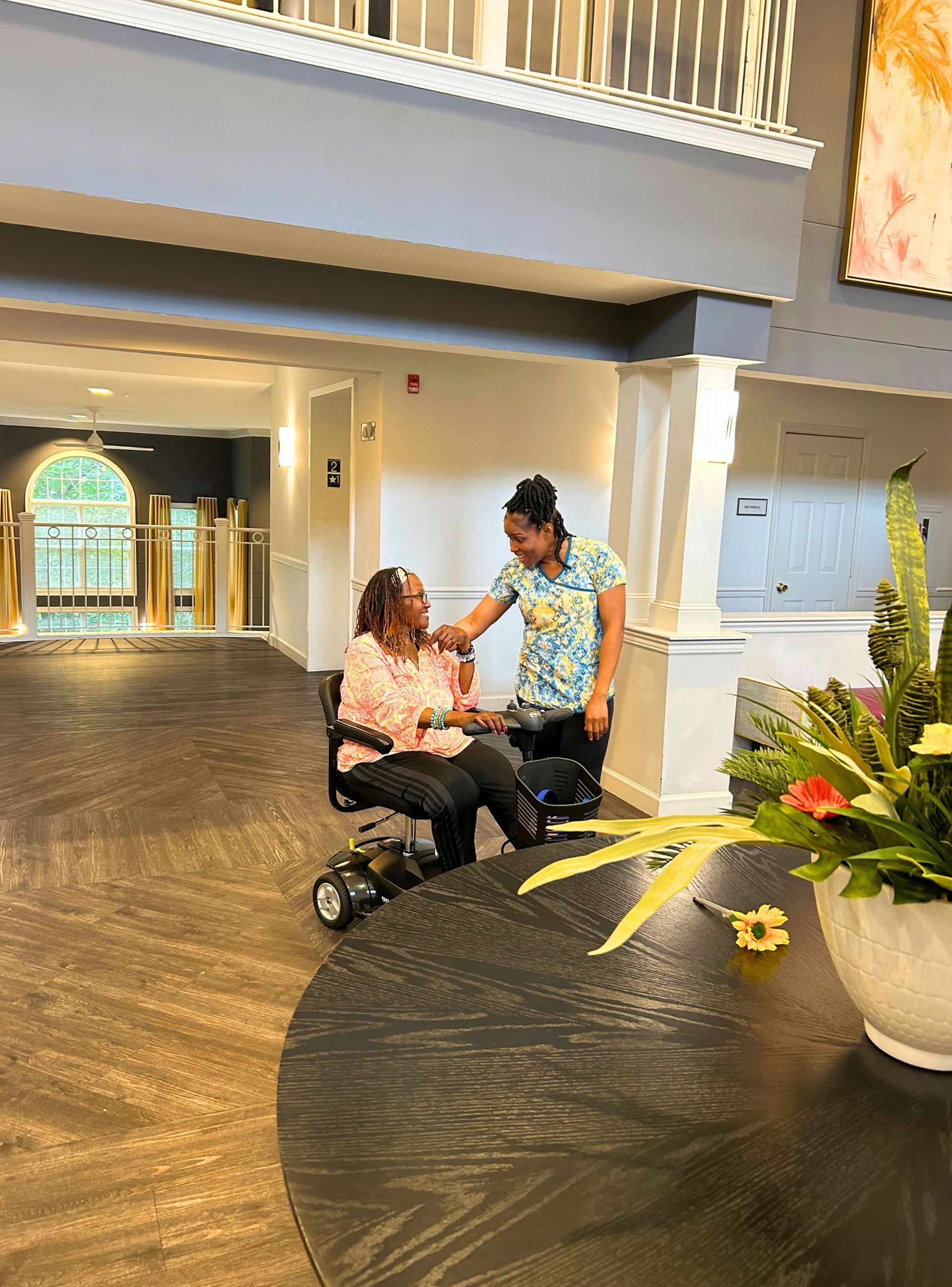 Woman in wheelchair smiles at caregiver in a hallway. Both are touching hands; light and airy interior.