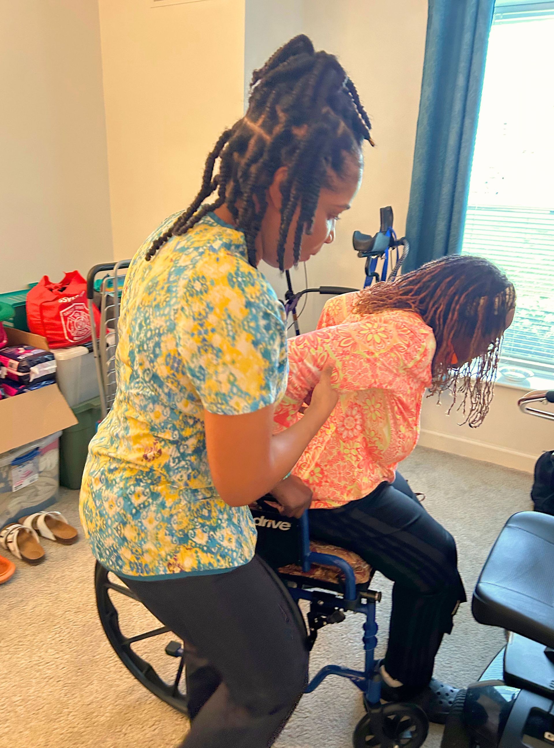 Woman assists another person from a wheelchair. Both have long hair, indoors by a window.