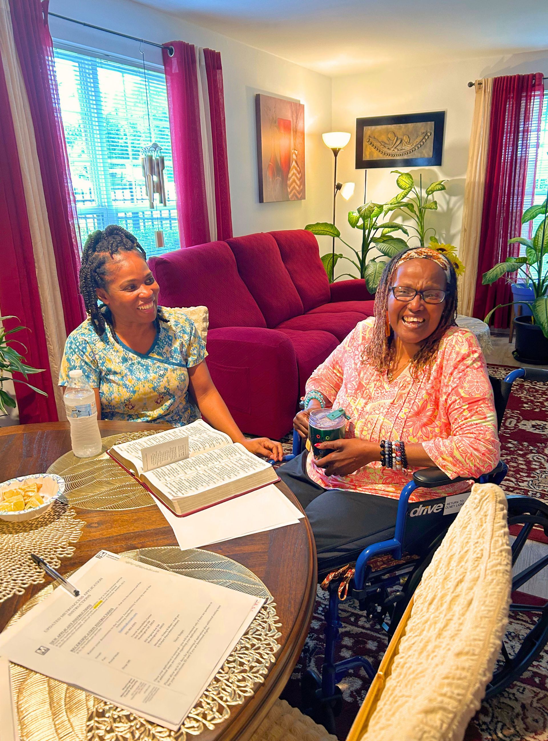 Two women sit at a table in a living room. One is in a wheelchair. Both are smiling.