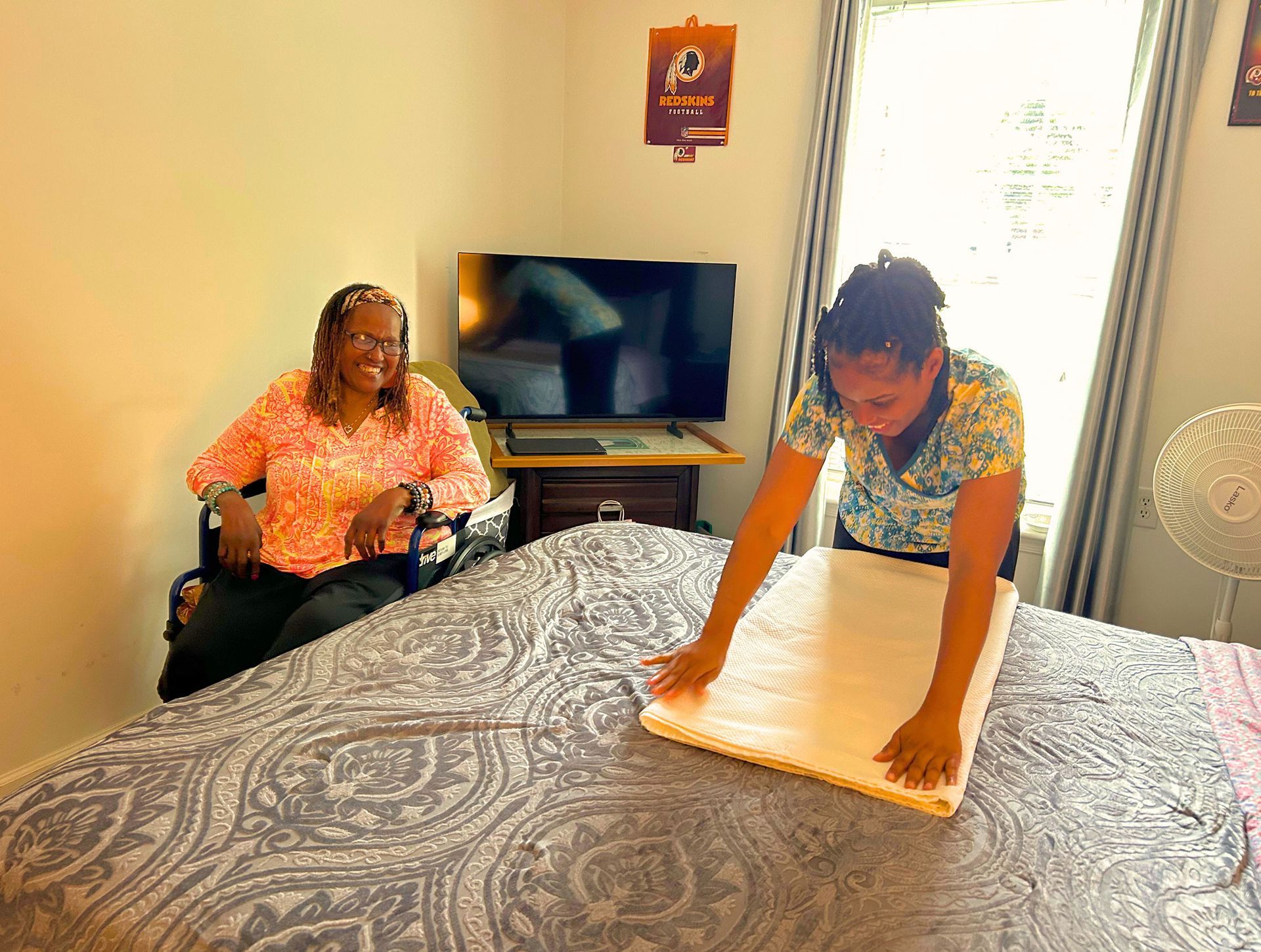 Two women in a bedroom. One smiles, sitting. The other smooths a mattress pad on a bed.