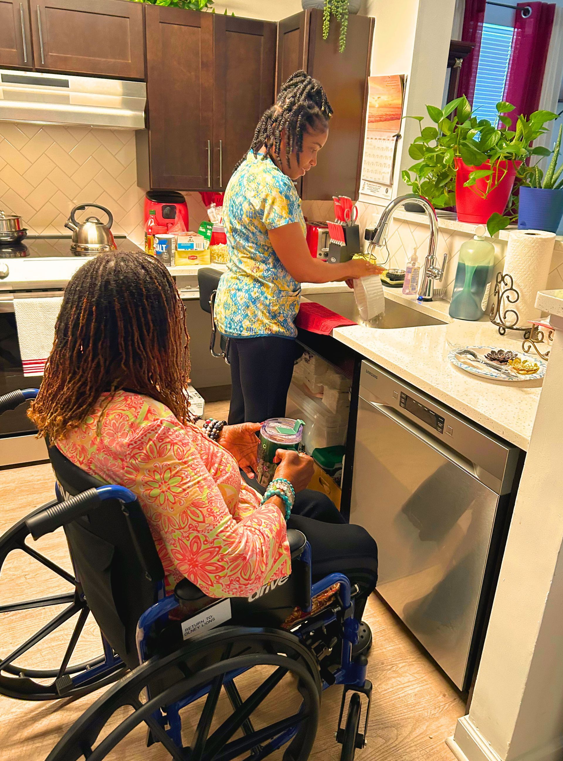 Woman in wheelchair watches another wash dishes in a kitchen.