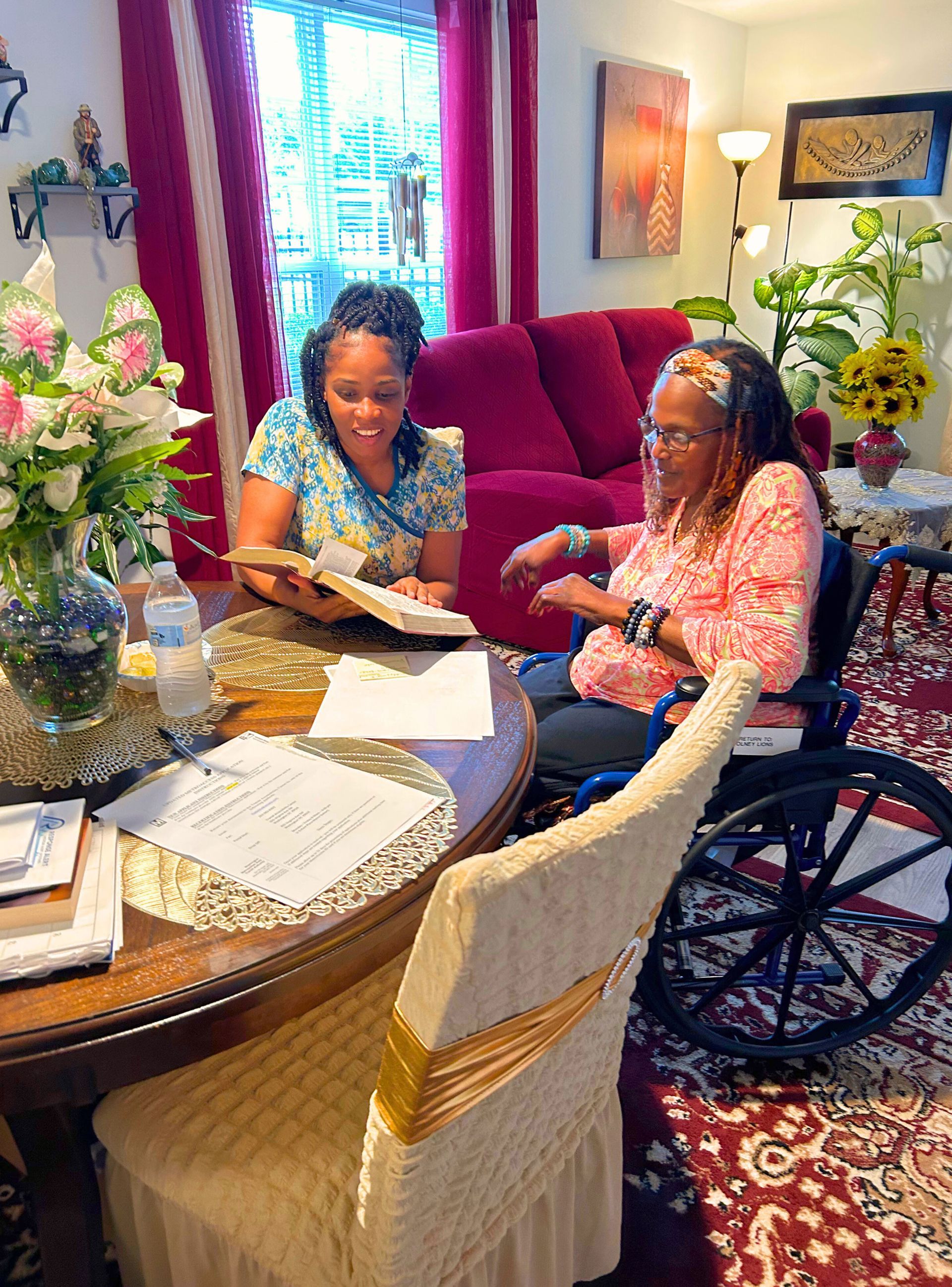 Two women seated at a table, one in a wheelchair, reading a book together in a living room.