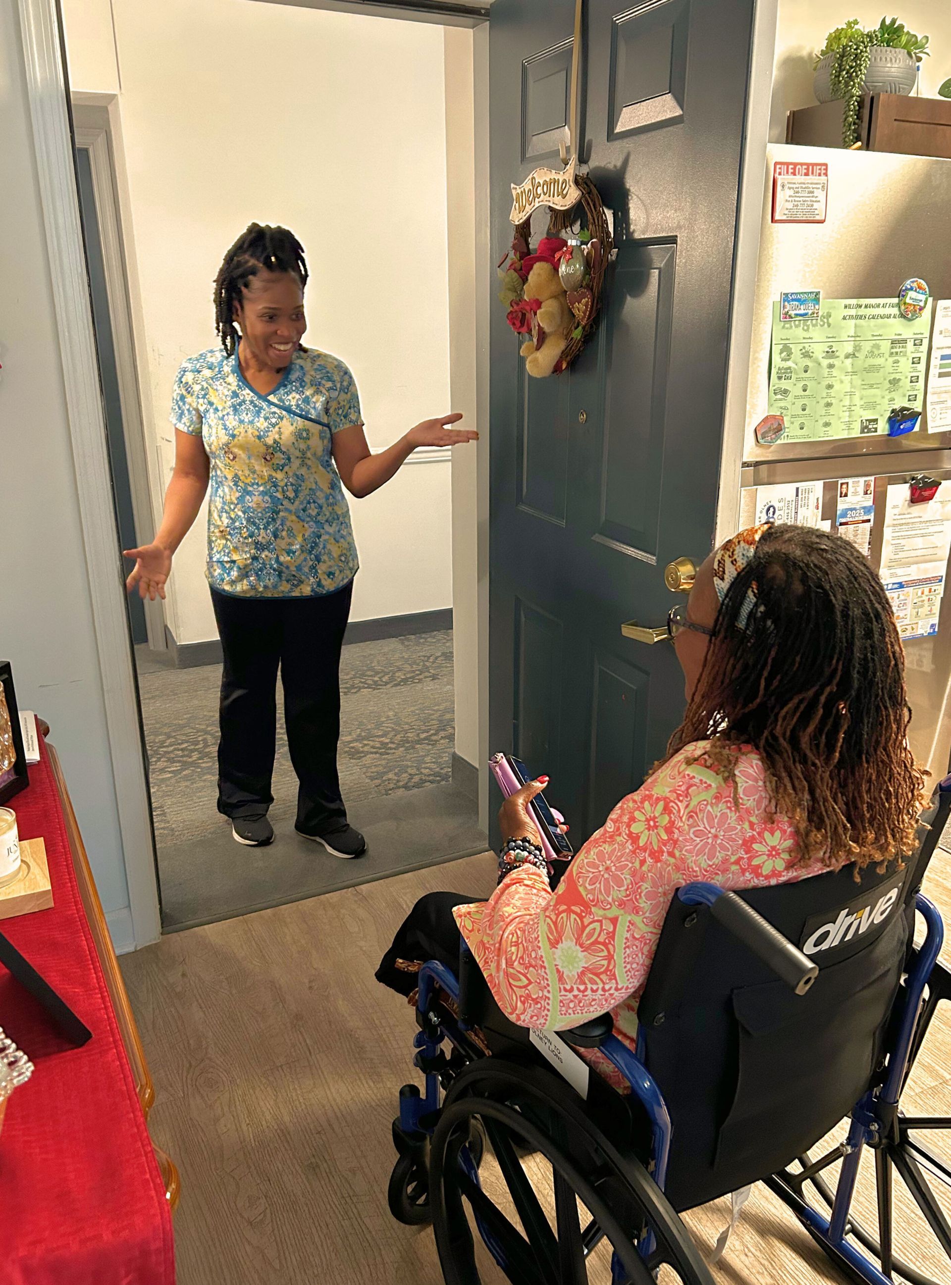 Woman in wheelchair greeted by another at doorway.