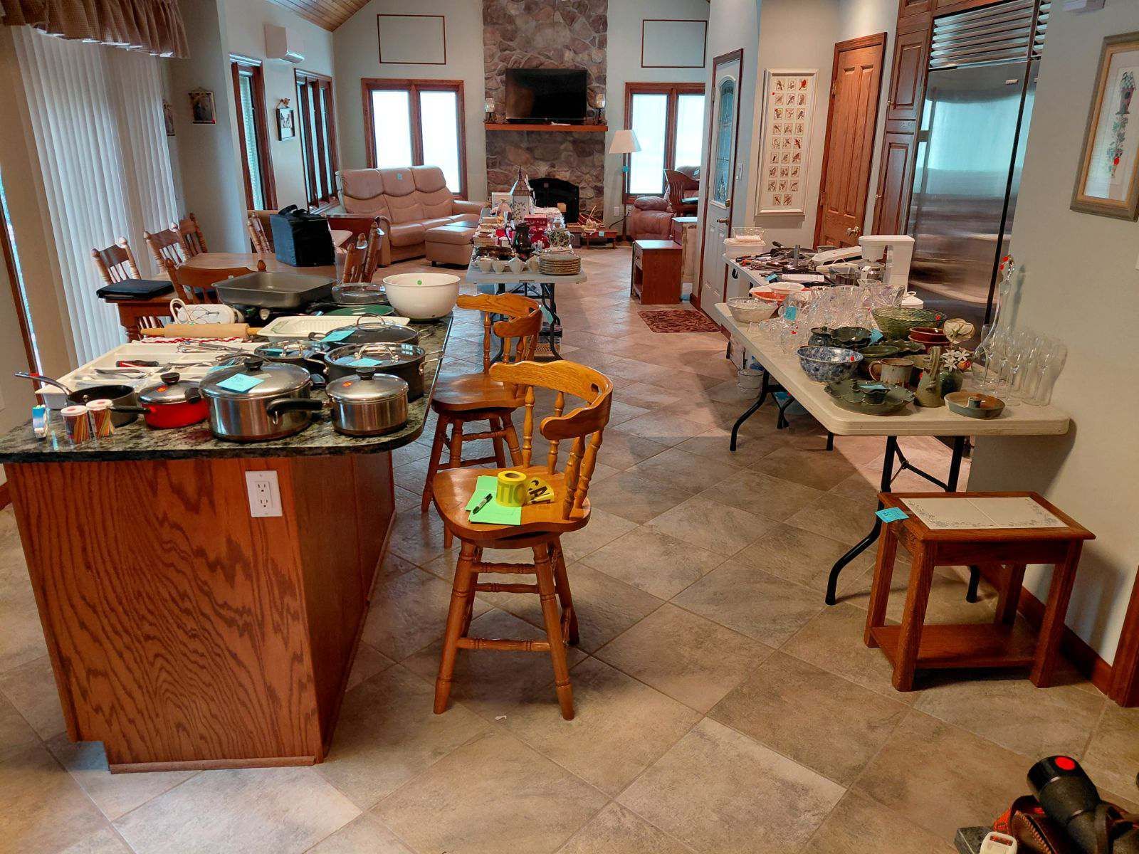 Kitchen/living room cluttered with items for sale, tables, island, brown cabinets, tan floor, open space, sunlight.