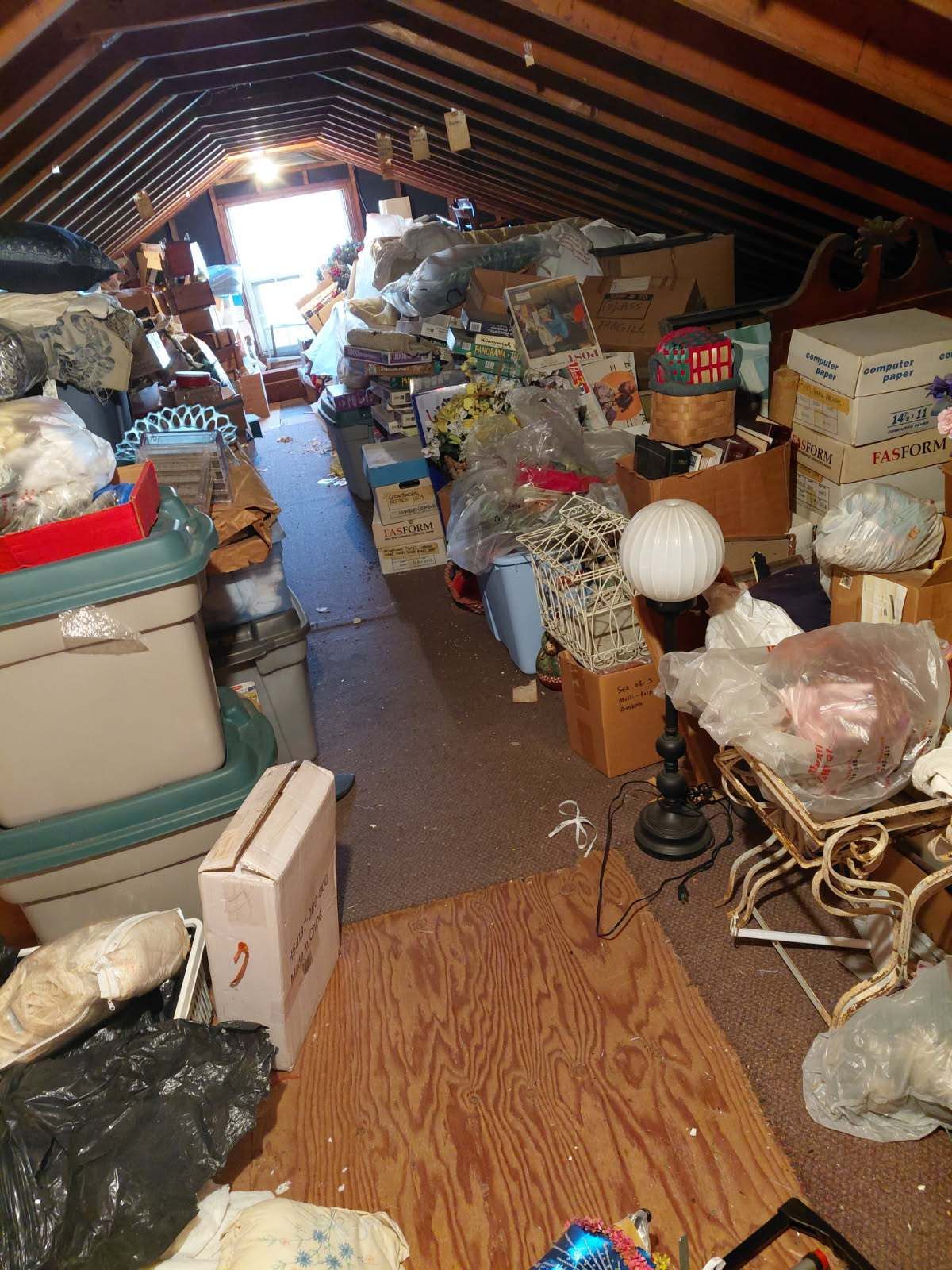 Cluttered attic filled with boxes, bins, and miscellaneous items; narrow walkway leads to a window.