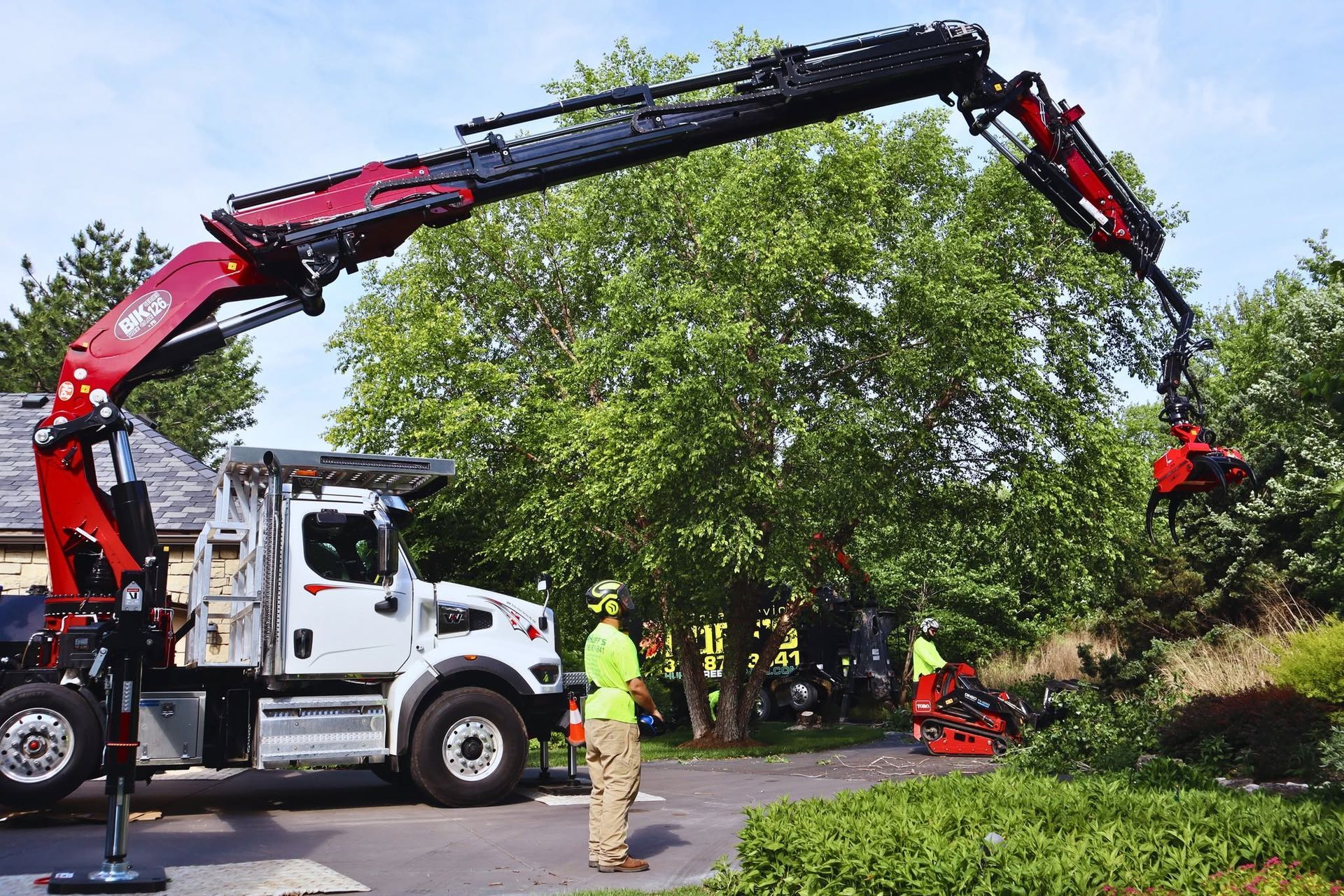 Tree-trimming truck with extended crane over a large green tree, worker standing nearby on a street