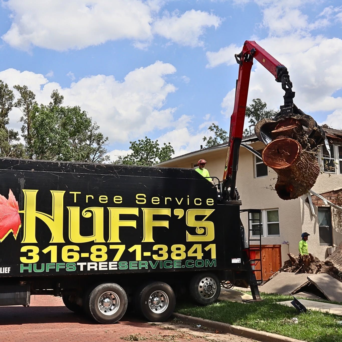 Tree service truck with crane lifting a large tree stump beside a house on a sunny day