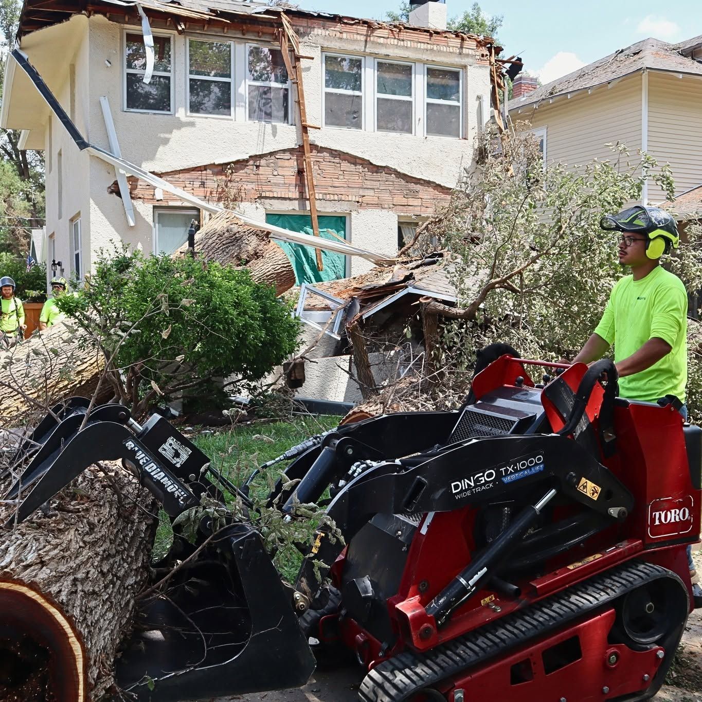 Worker in neon shirt operating a red skid-steer amid debris in front of a damaged house.
