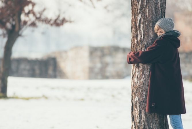 Person in a gray hat and dark coat hugging a tree in a snowy park