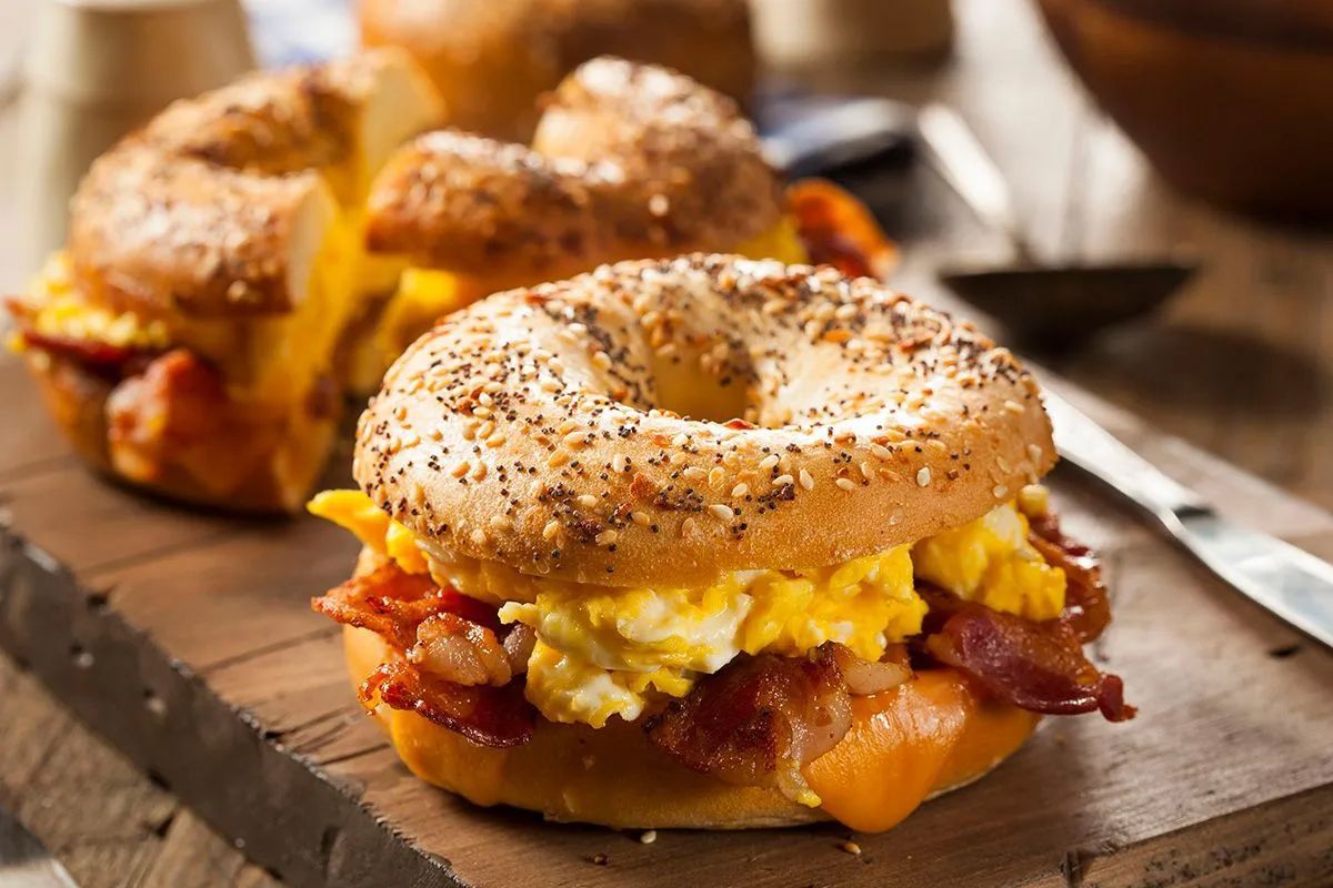 A bagel with eggs, bacon, and cheese on a wooden cutting board.
