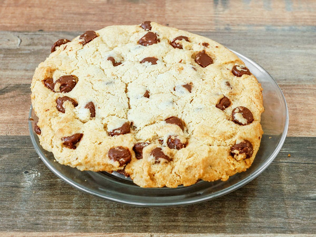 A chocolate chip cookie is on a glass plate on a wooden table.