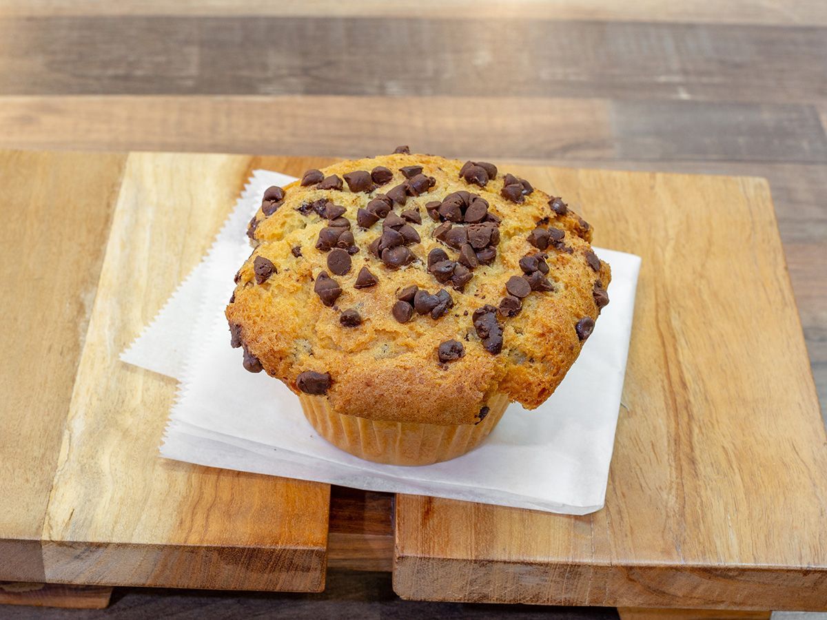 A chocolate chip muffin is sitting on a napkin on a wooden cutting board.
