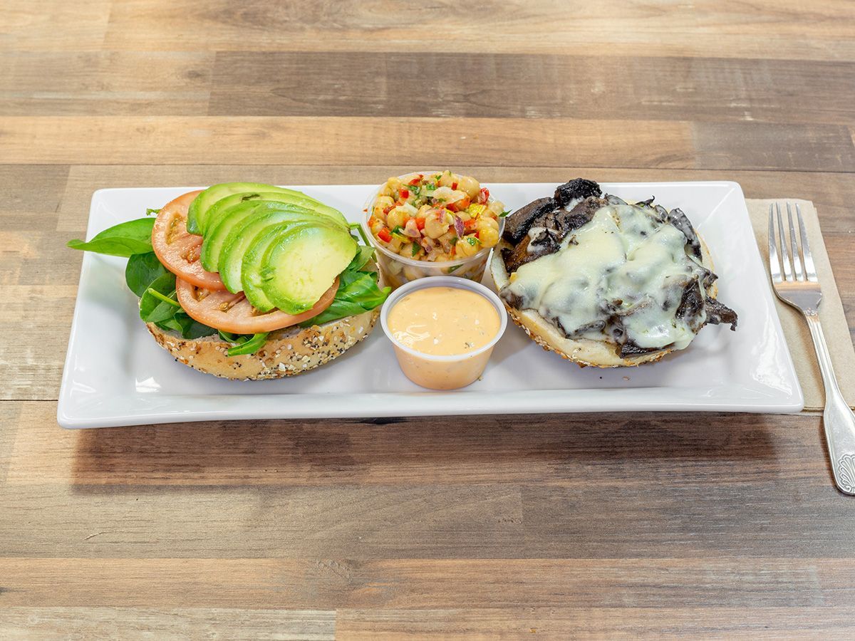 A bagel sandwich with avocado , tomato, and mushrooms on a white plate on a wooden table.