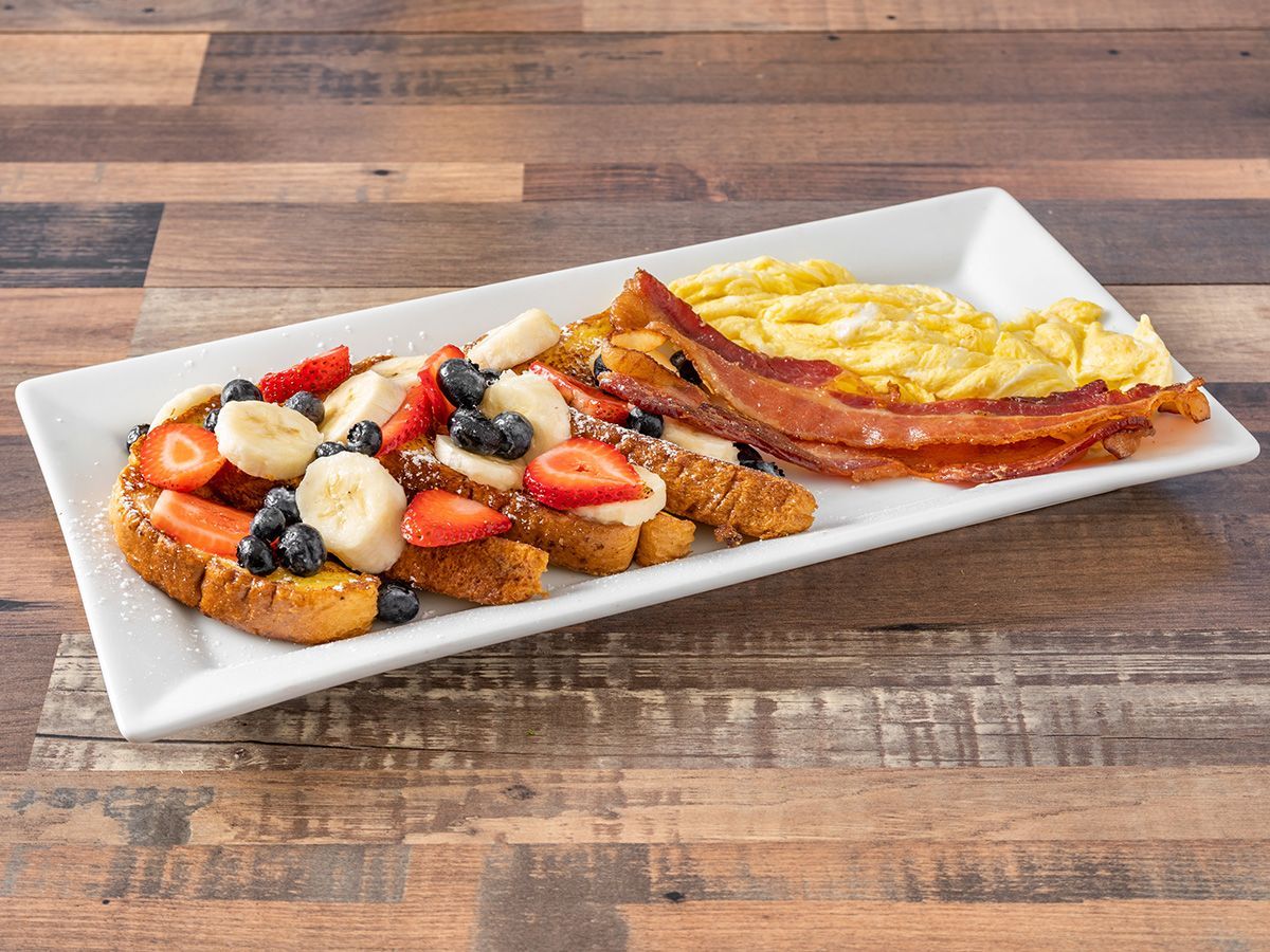 A white plate topped with french toast, eggs, bacon and fruit on a wooden table.