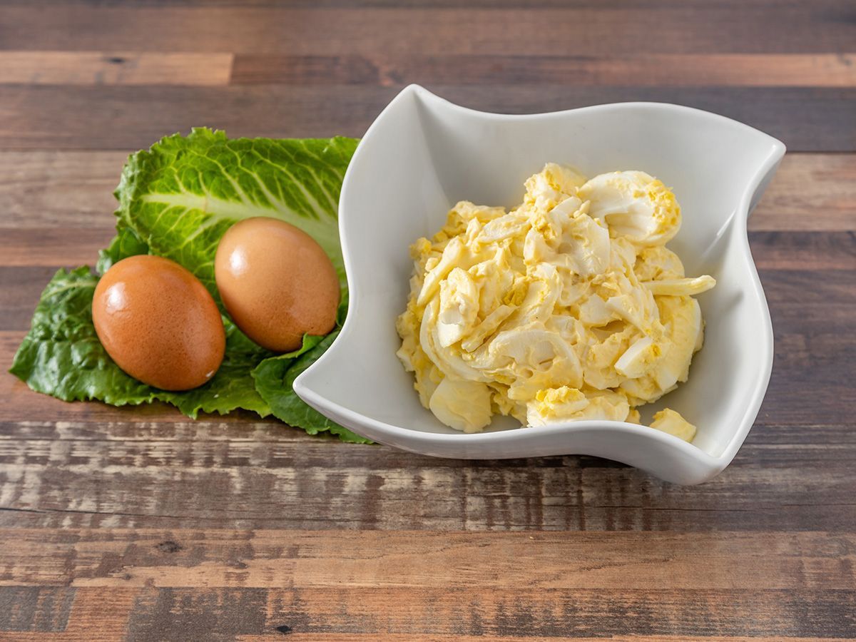 A bowl of scrambled eggs next to two eggs and lettuce on a wooden table.