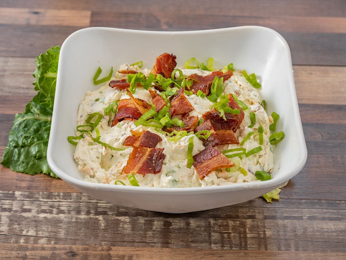A bowl of potato salad with bacon and green onions on a wooden table.