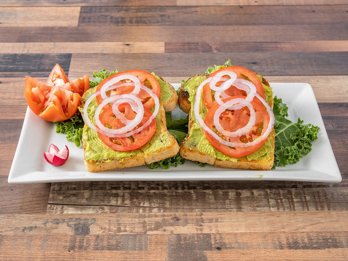 Two slices of avocado toast with tomatoes and onions on a white plate on a wooden table.