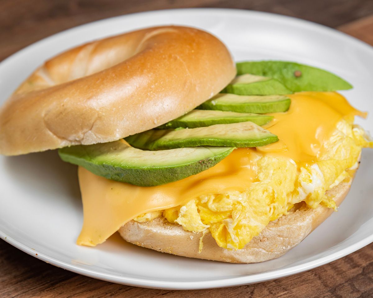 A bagel sandwich with eggs, cheese and avocado on a white plate.