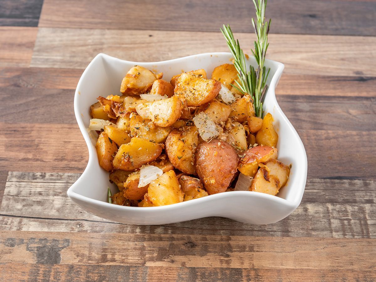 A bowl of fried potatoes with a sprig of rosemary on a wooden table.