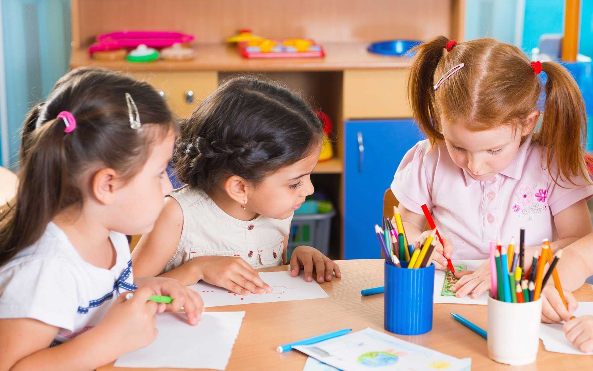 Three little girls are sitting at a table drawing with colored pencils.