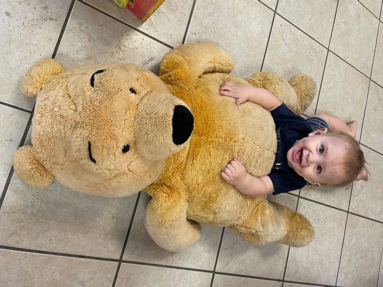 Smiling child lying on a large tan teddy bear on a tiled floor