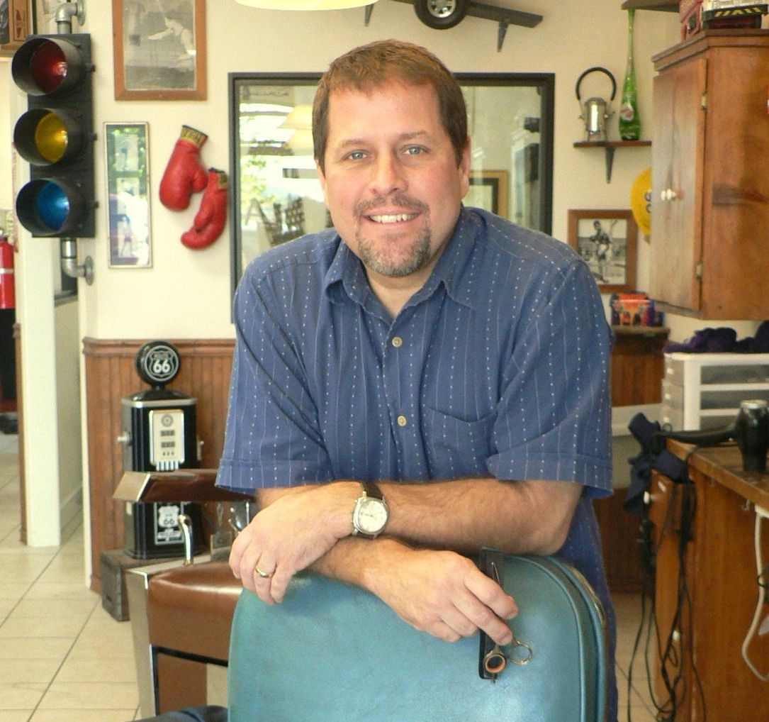 Man smiling in a barber shop, leaning on a chair with vintage decor and a traffic light in the background