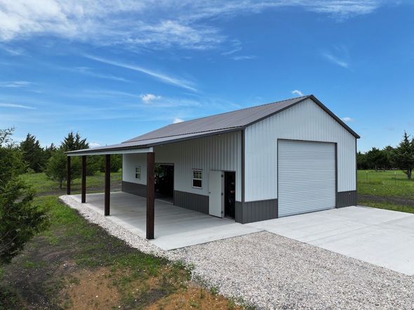 Metal-sided building with a carport, gray roof, and large garage door. Concrete driveway and gravel approach.
