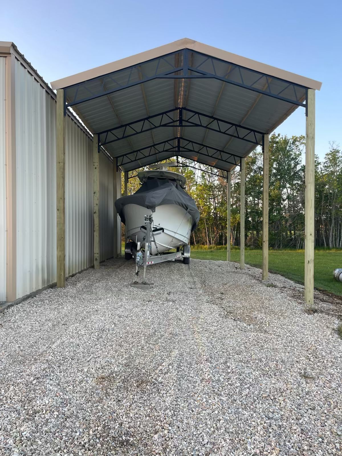Boat on a trailer sheltered under a metal roof attached to a building and wooden posts, on a gravel driveway.