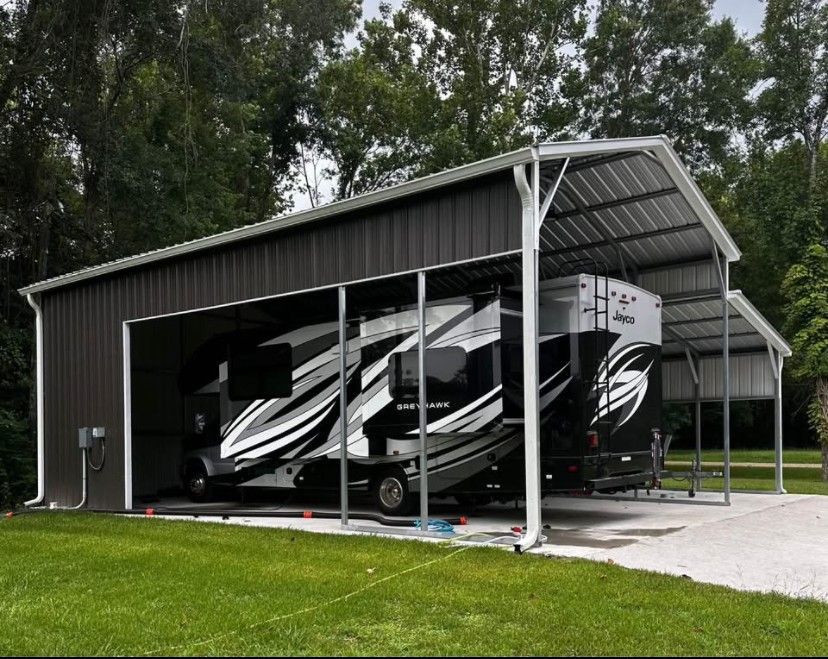 RV parked under a dark brown metal carport on green grass, with trees in the background.