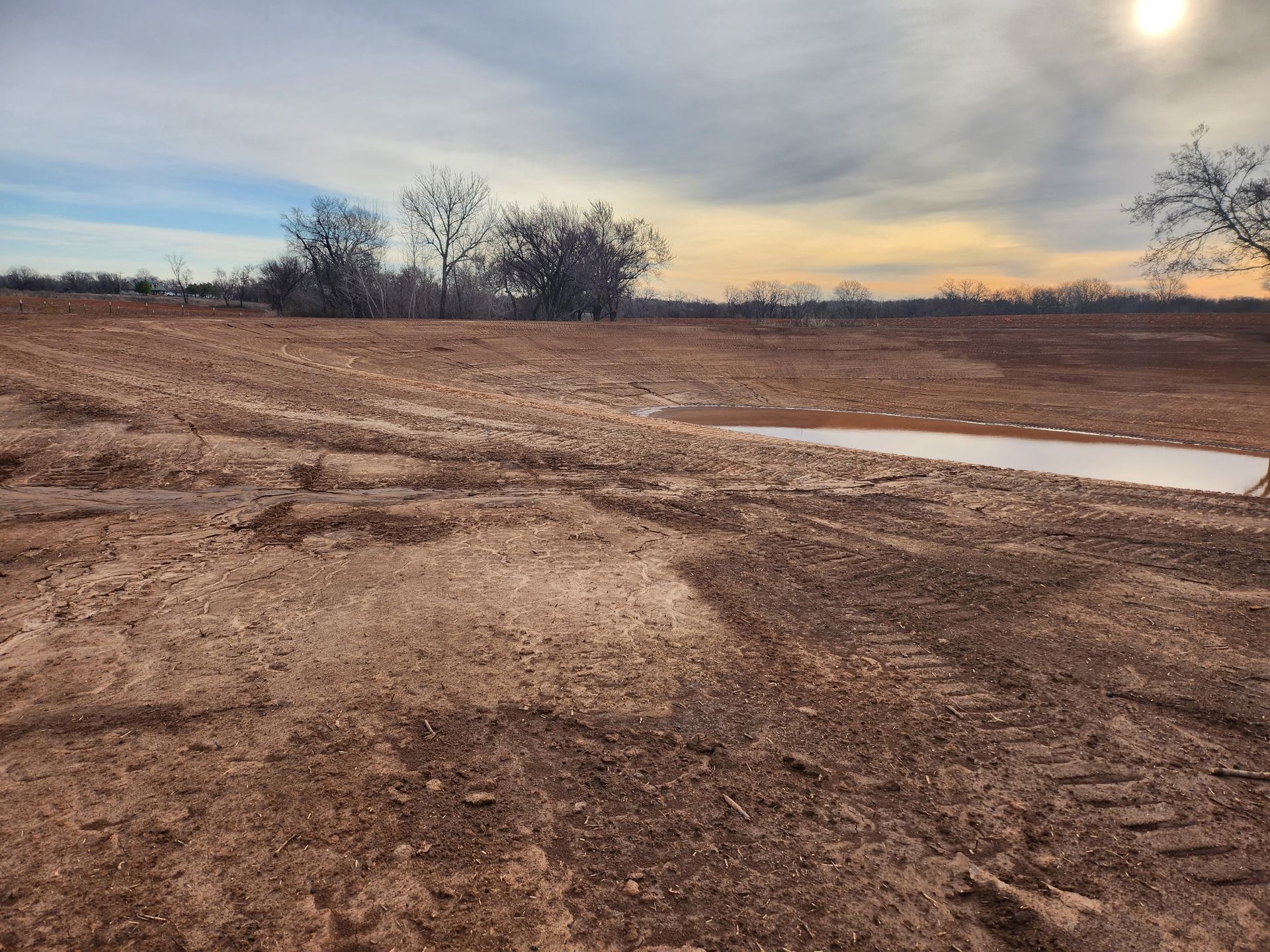 A dirt field with trees in the background and the sun shining through the clouds