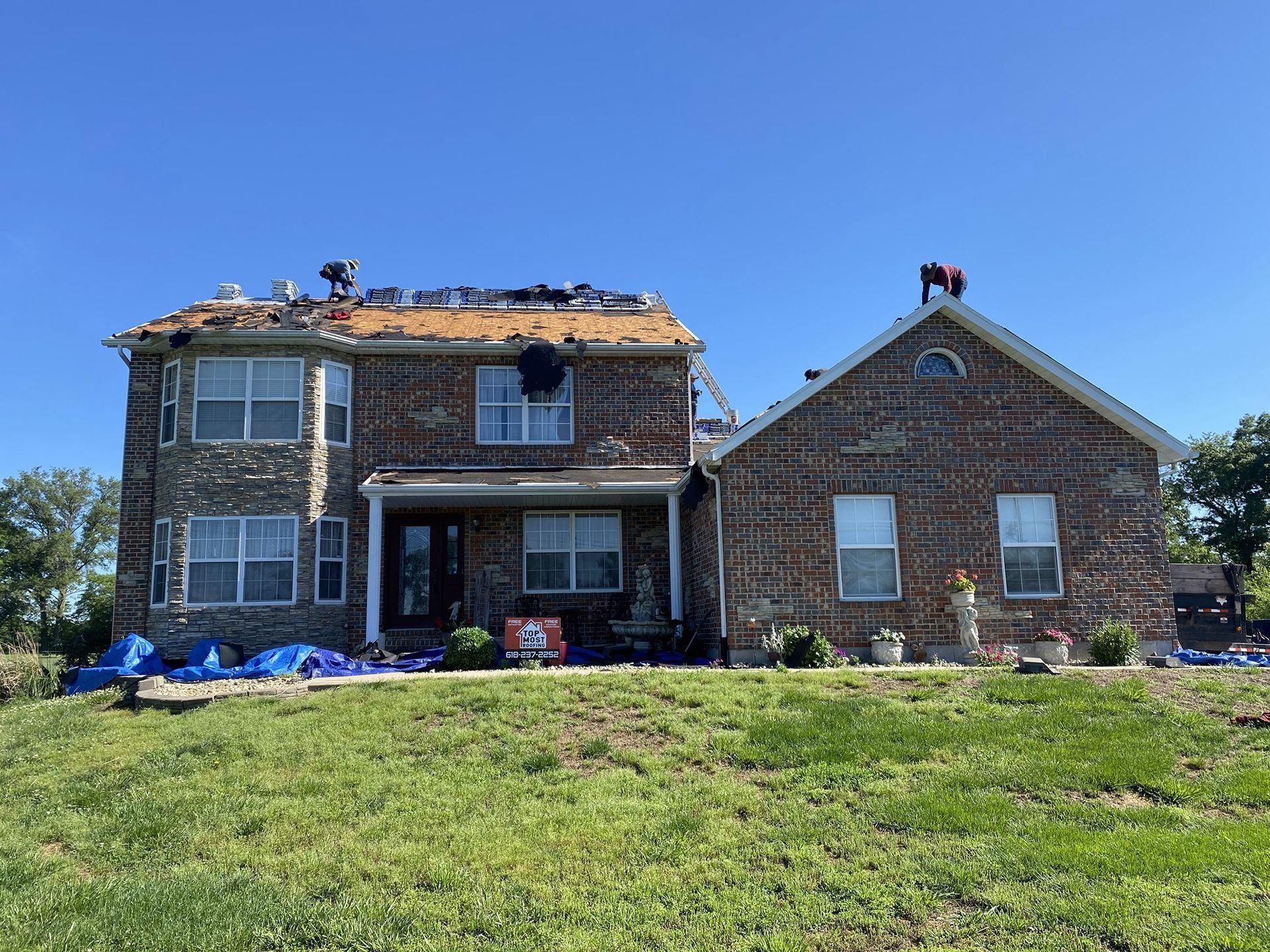 Damaged house with roof torn off, debris, and a blue tarp on the lawn under a clear blue sky