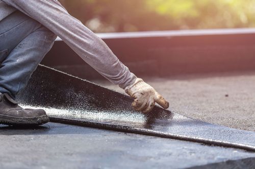 Worker laying black roofing material on a rooftop with a trowel and gloves