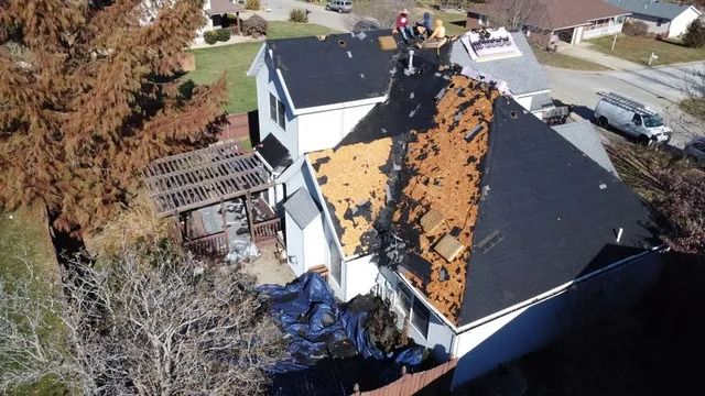 Aerial view of a house with severe roof damage, missing shingles, and debris near neighboring homes.