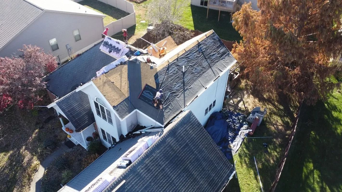 Aerial view of a house with roof damage and debris, surrounded by trees and neighboring homes.