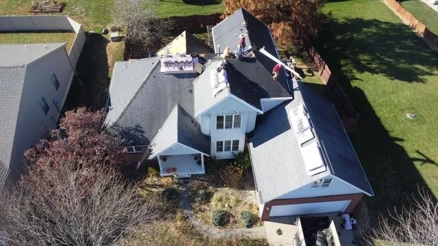 Aerial view of a gray-roofed house with dormers, chimneys, and a front porch surrounded by a yard.