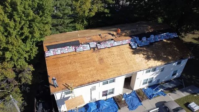 Aerial view of a roof under repair, with exposed plywood, tarps, and a few workers near the edge.