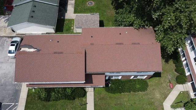 Aerial view of a single-story house with a red roof, green lawn, driveway, and trees around it.